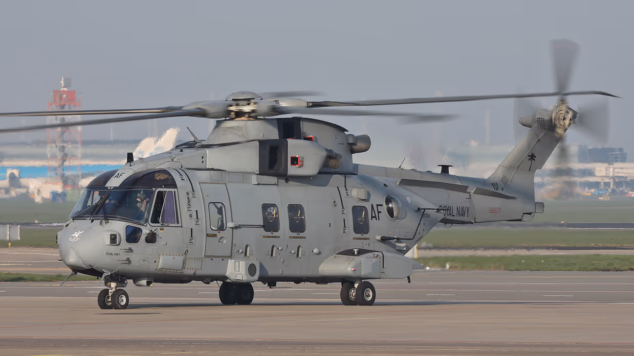  Een grote, grijze militaire helikopter met draaiende rotorbladen staat op een startbaan, met "Amsterdam" en taxiborden op de achtergrond.