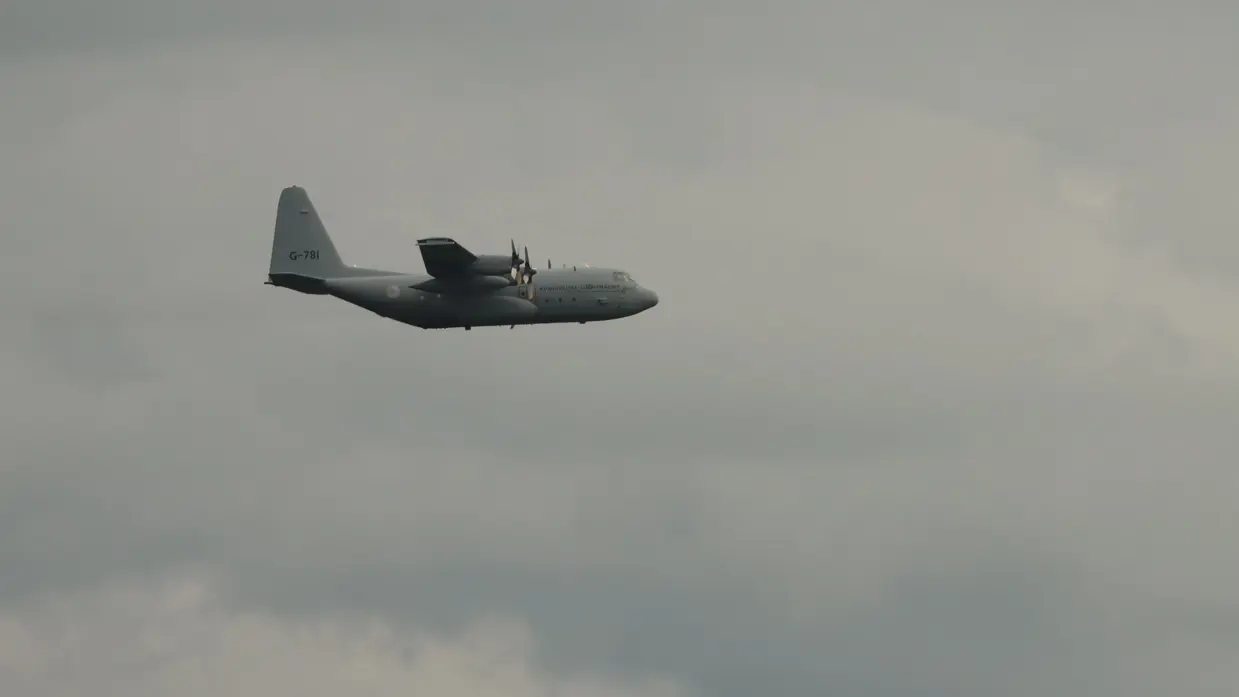  Een grijs militair vliegtuig vliegt onder een blauwe hemel over een dak van een huis.
