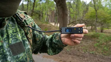 Links: iemand trekt een groene bergschoen aan met een zwarte beugel, buiten op een bosbodem met bladeren.    Midden: Iemand in camouflagebroek zit op een auto met bruine sokken en houdt een groene bergschoen vast. Rechts: Een man in uniform houdt een metertje vast. 