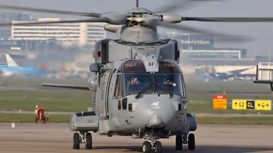  Een grote, grijze militaire helikopter met draaiende rotorbladen staat op een startbaan, met "Amsterdam" en taxiborden op de achtergrond.