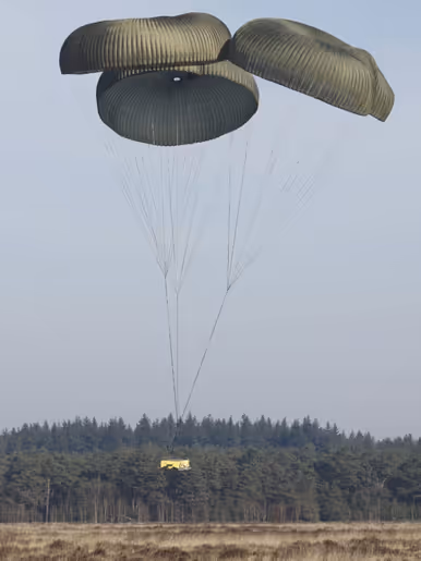 Een C-130 in de lucht met vlak achter het toestel een groot pakket met daaraan een horizontaal gespannen lijn met aan de andere kant een parachute.