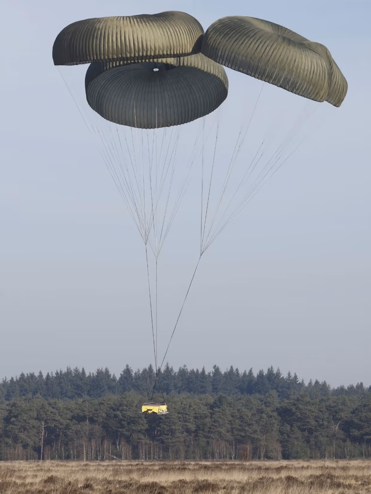 Een C-130 in de lucht met vlak achter het toestel een groot pakket met daaraan een horizontaal gespannen lijn met aan de andere kant een parachute.