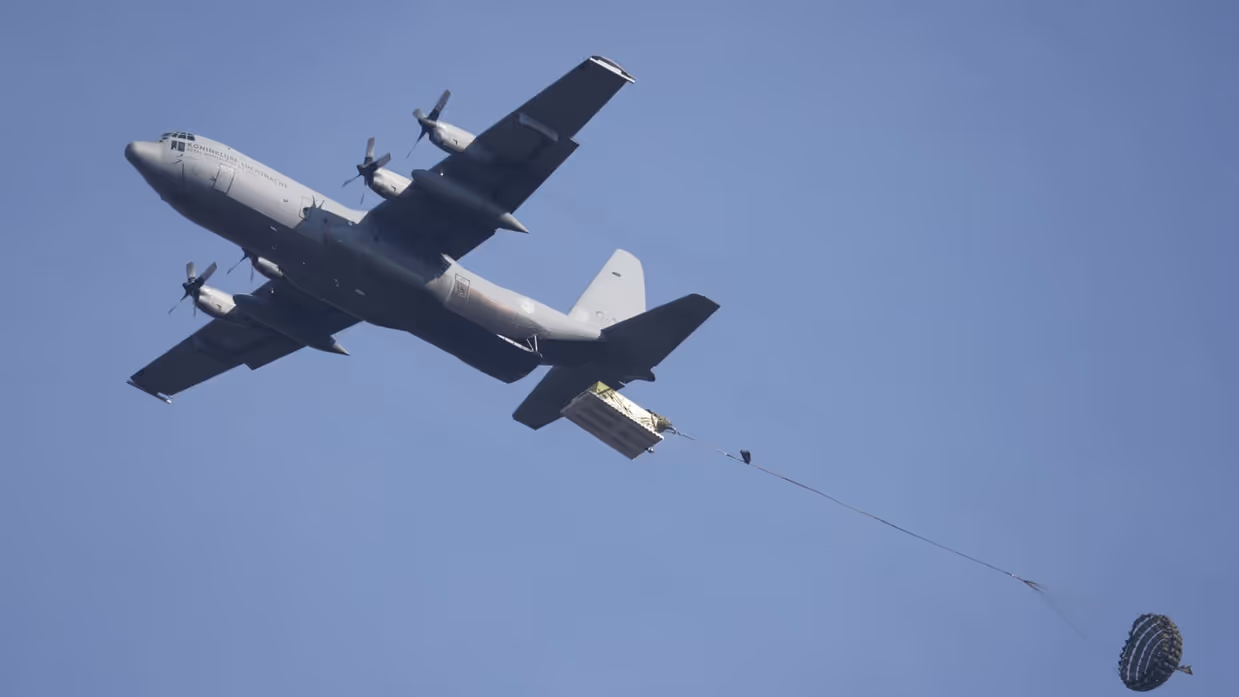 Een C-130 in de lucht met vlak achter het toestel een groot pakket met daaraan een horizontaal gespannen lijn met aan de andere kant een parachute.