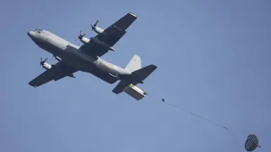 Een C-130 in de lucht met vlak achter het toestel een groot pakket met daaraan een horizontaal gespannen lijn met aan de andere kant een parachute.