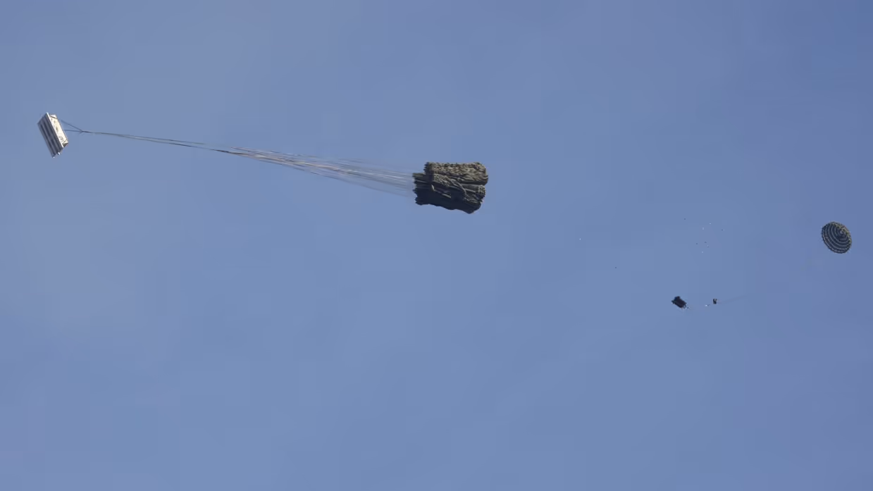 Een C-130 in de lucht met vlak achter het toestel een groot pakket met daaraan een horizontaal gespannen lijn met aan de andere kant een parachute.