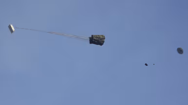 Een C-130 in de lucht met vlak achter het toestel een groot pakket met daaraan een horizontaal gespannen lijn met aan de andere kant een parachute.