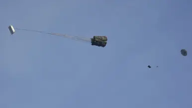 Een C-130 in de lucht met vlak achter het toestel een groot pakket met daaraan een horizontaal gespannen lijn met aan de andere kant een parachute.