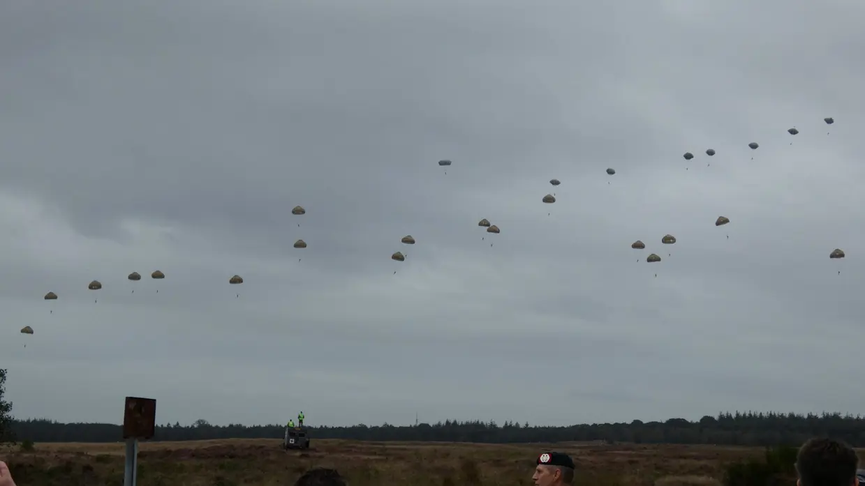  Meerdere para's dalen uit de lucht boven een open veld, toeschouwers kijken toe onder een bewolkte hemel. 