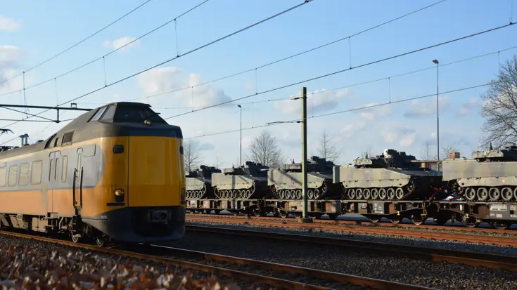 Een geel-blauwe trein van de Nederlandse Spoorwegen rijdt langs wagons militaire pantservoertuigen op een spoor, met bovenleidingen en een blauwe hemel. 