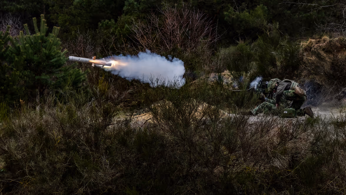 Een soldaat in camouflagekleding schiet in een bosrijke omgeving een raket af met zichtbare rook en vuur uit de achterkant.