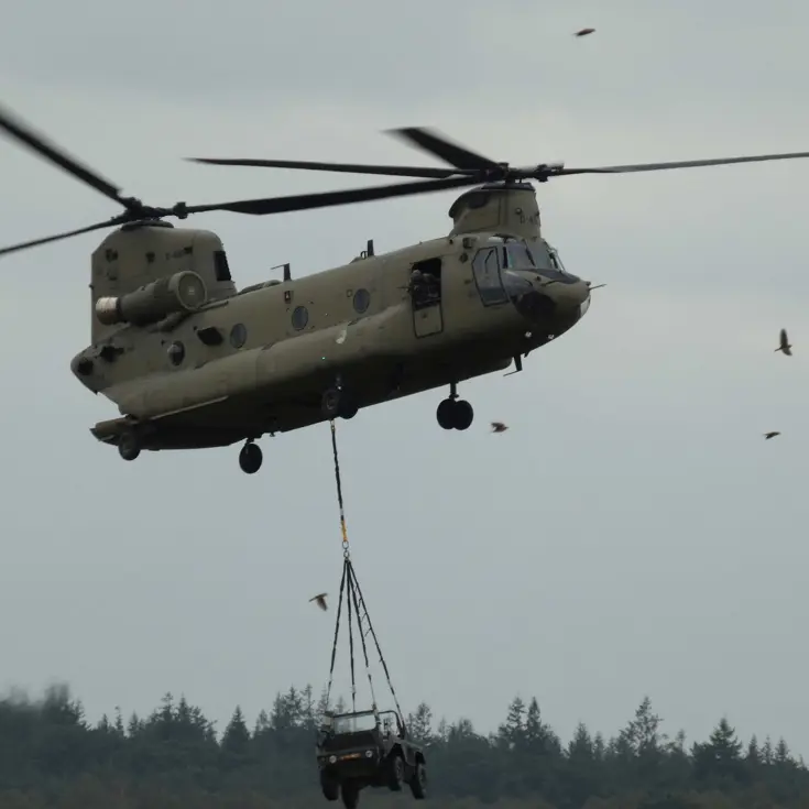 Een militaire helikopter tilt een terreinvoertuig op onder een bewolkte hemel, met bomen en vogels op de achtergrond.