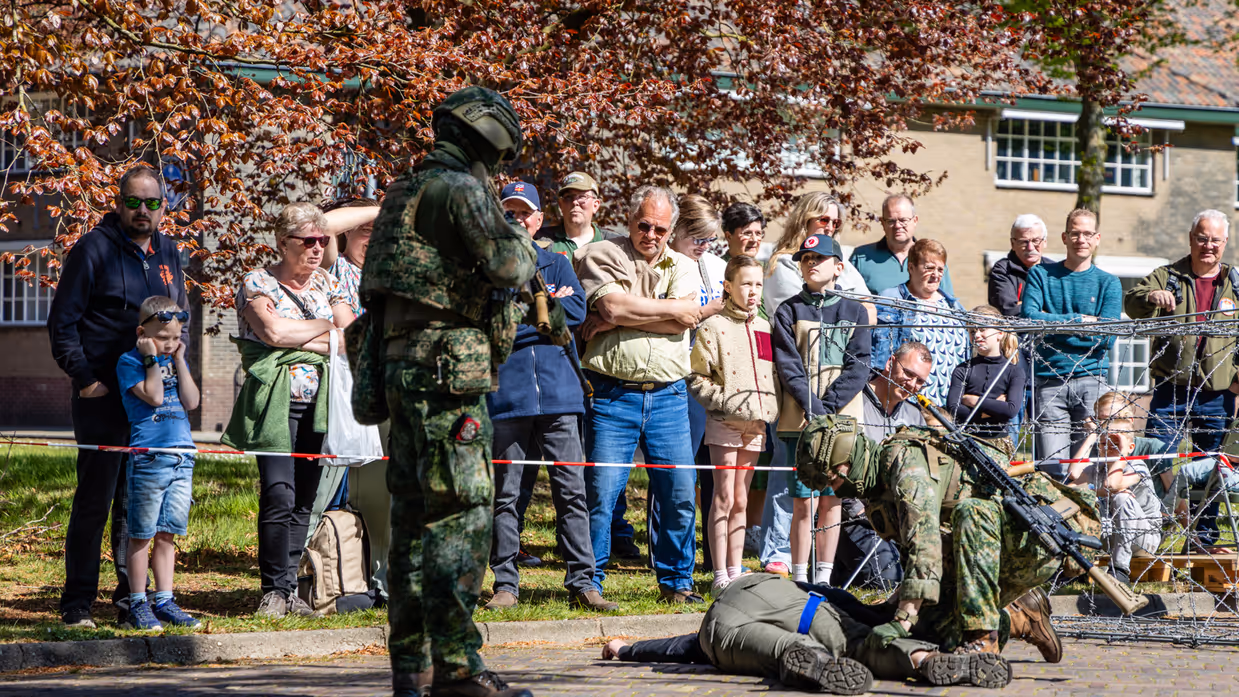 Soldaten in camouflage voeren een oefening uit, omringd door toeschouwers achter prikkeldraad en bomen. 