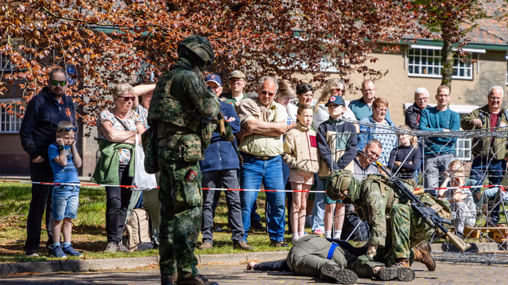Soldaten in camouflage voeren een oefening uit, omringd door toeschouwers achter prikkeldraad en bomen. 