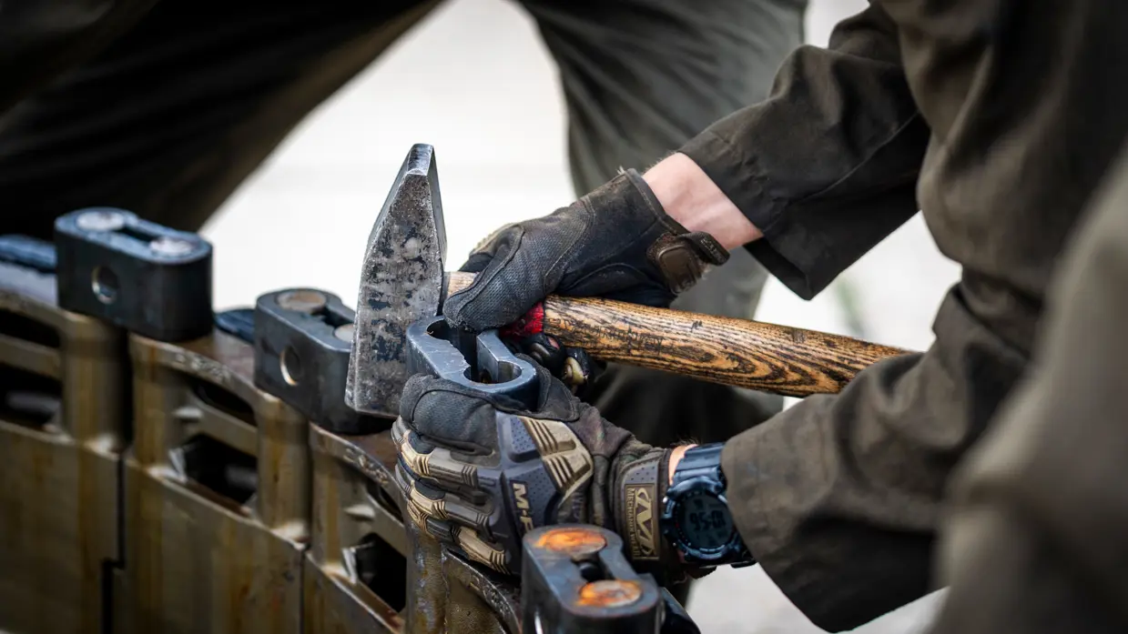Handen in beschermende handschoenen gebruiken een hamer en een houten steel om een metalen onderdeel te bewerken.
