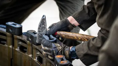 Handen in beschermende handschoenen gebruiken een hamer en een houten steel om een metalen onderdeel te bewerken.