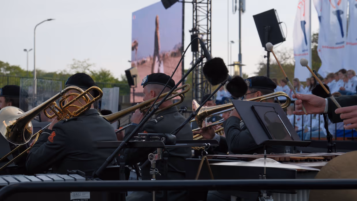 Een buitenoptreden: brassband met trompetten en trombones, microfoons, bladmuziek, publiek op achtergrond, avondlicht en vlaggen. 