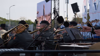 Een buitenoptreden: brassband met trompetten en trombones, microfoons, bladmuziek, publiek op achtergrond, avondlicht en vlaggen. 