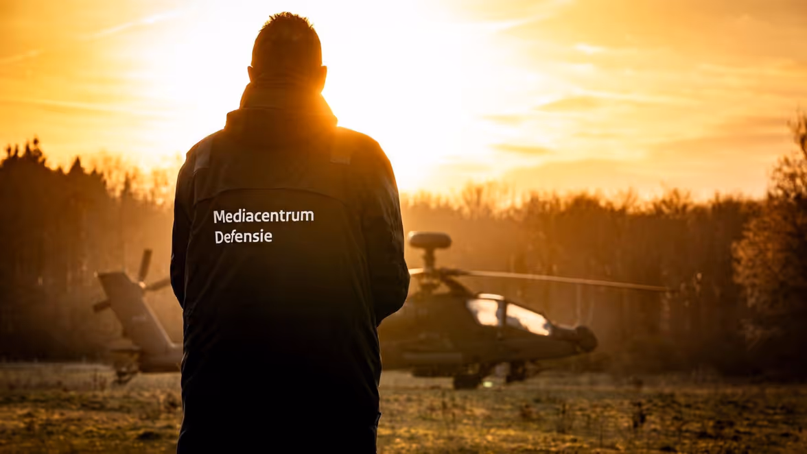 Een persoon van achteren, gekleed in een jas met "Mediacentrum Defensie" erop, staand voor een helikopter tijdens een zonsondergang in een natuurlijke omgeving.