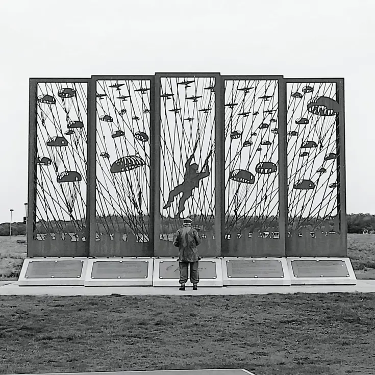 Het silhouet van een man staat voor een groot metalen monument met parachutisten en vliegtuigen, in een open veld.