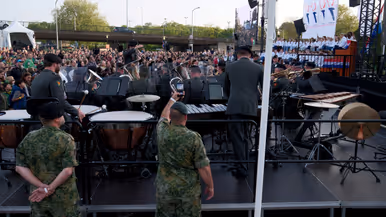 Groot buitenconcert: militaire band speelt op podium, publiek kijkt toe, soldaten in camouflage, drums, trompetten, vlaggen en bomen. 