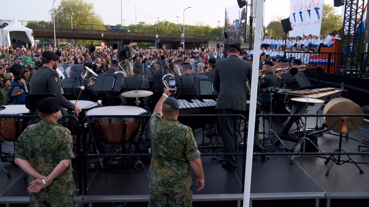 Groot buitenconcert: militaire band speelt op podium, publiek kijkt toe, soldaten in camouflage, drums, trompetten, vlaggen en bomen. 