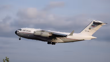 Een wit vliegtuig met de tekst "KUWAIT AIR FORCE" en nummer "KAF 343" vliegt door een bewolkte lucht. 