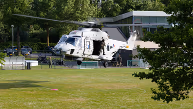 Een witte militaire helikopter met open deur, twee bemanningsleden, staat op een grasveld bij een gebouw en bomen. 