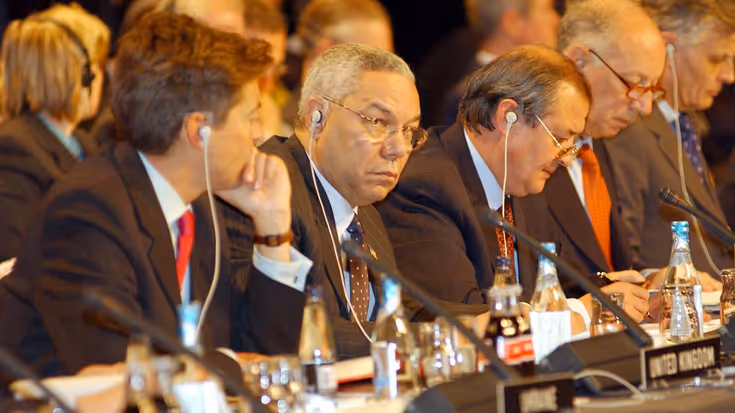 Een groep mensen in pak tijdens een officiële vergadering. Ze zitten aan tafels met microfoons, naamplaatjes, koptelefoons en drinkflesjes, en zien er geconcentreerd uit.