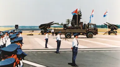 Militairen in donkerblauwe broek en lichtblauwe bloes staan op de plaats rust. In de achtergrond een Patriot- en twee HAWK-lanceerinrichtingen, een vrachtwagen en drie vlaggen.