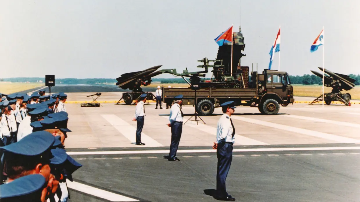 Militairen in donkerblauwe broek en lichtblauwe bloes staan op de plaats rust. In de achtergrond een Patriot- en twee HAWK-lanceerinrichtingen, een vrachtwagen en drie vlaggen.