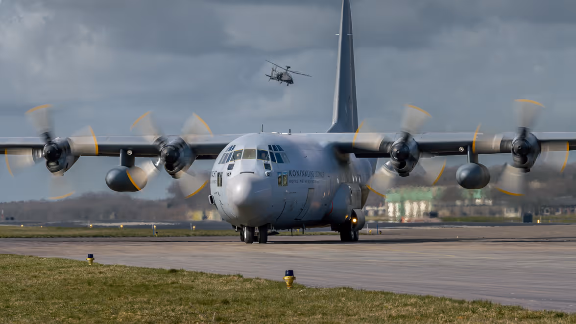 Een groot militair transportvliegtuig met vier draaiende propellers op een startbaan, terwijl een helikopter er boven vliegt tegen een bewolkte lucht.