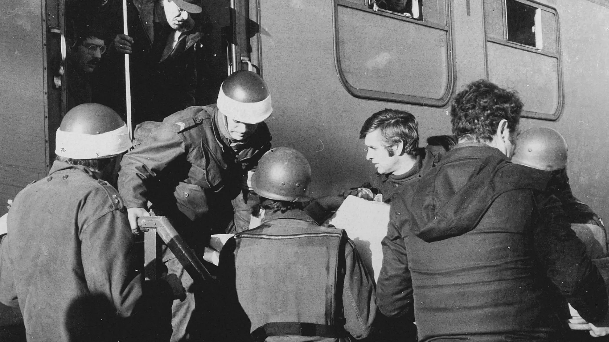 Deze zwart-wit foto toont militairen in gevechtsuitrusting die mensen helpen bij het uitstappen van een treinstel. Er is spanning zichtbaar. 