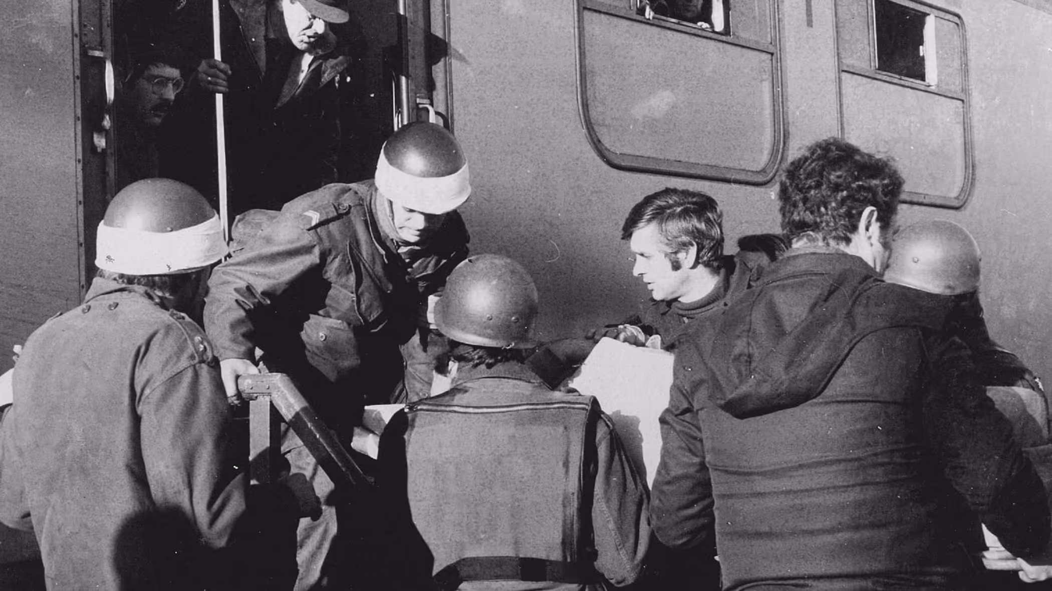 Deze zwart-wit foto toont militairen in gevechtsuitrusting die mensen helpen bij het uitstappen van een treinstel. Er is spanning zichtbaar. 