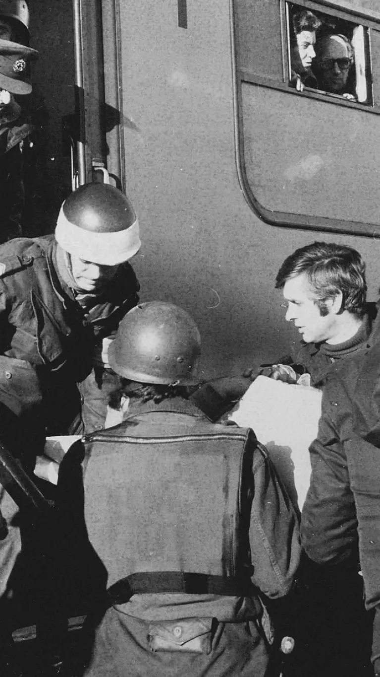 Deze zwart-wit foto toont militairen in gevechtsuitrusting die mensen helpen bij het uitstappen van een treinstel. Er is spanning zichtbaar. 