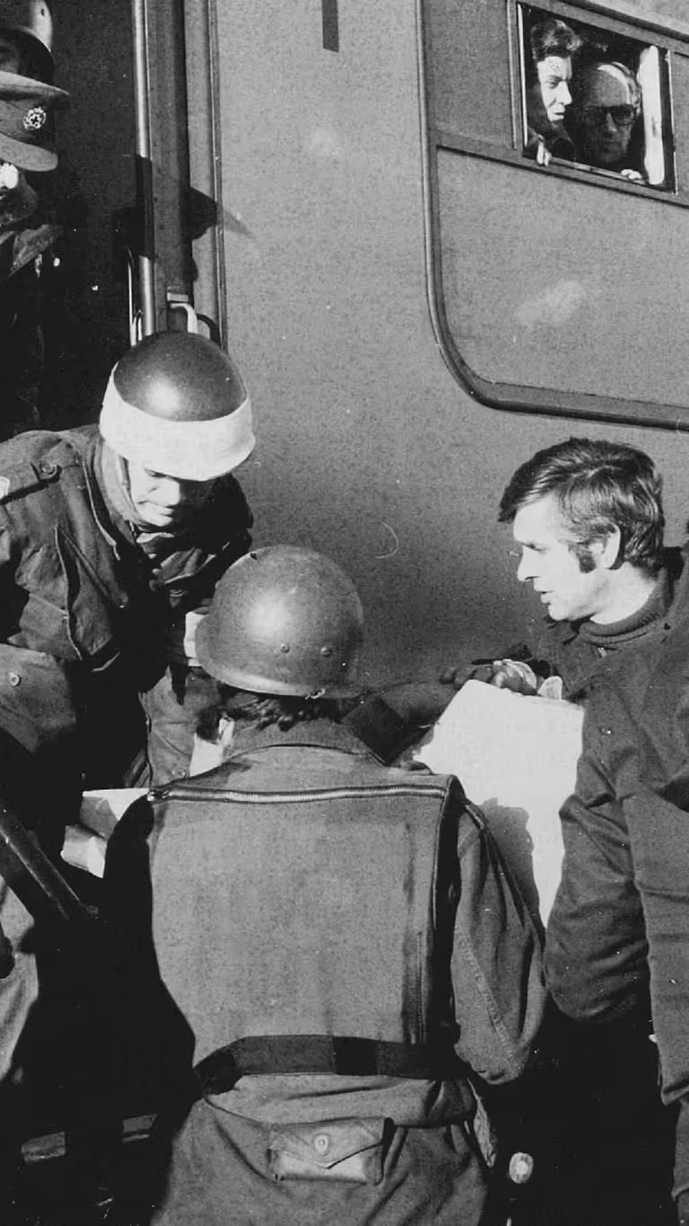 Deze zwart-wit foto toont militairen in gevechtsuitrusting die mensen helpen bij het uitstappen van een treinstel. Er is spanning zichtbaar. 