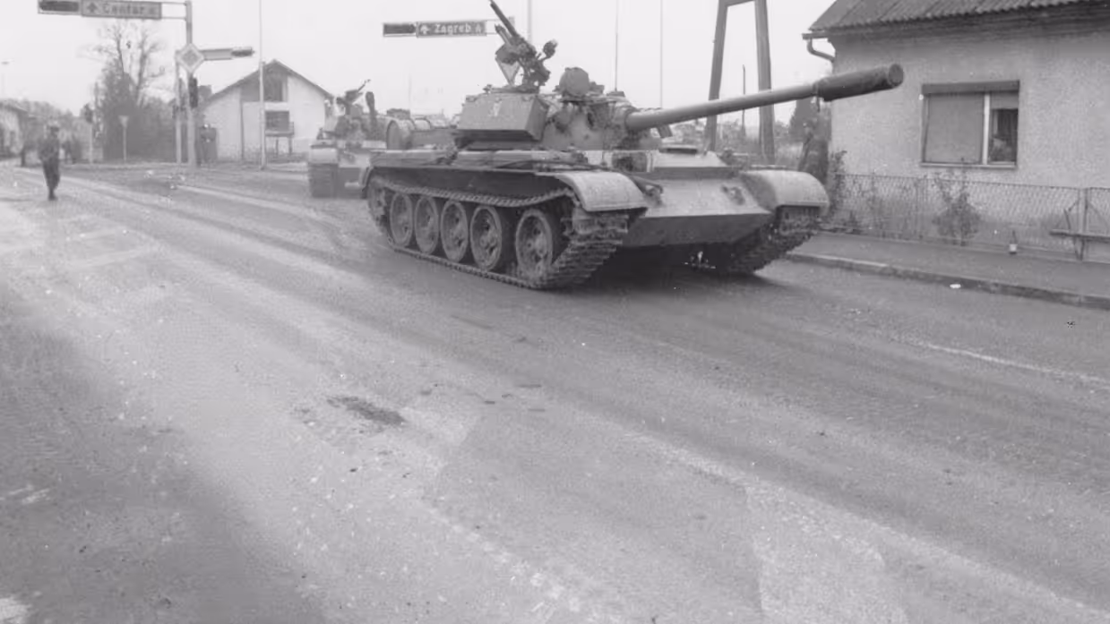 een tank rijdt langs een huis, met op de achtergrond een verkeersbord met daarop Zagreb.