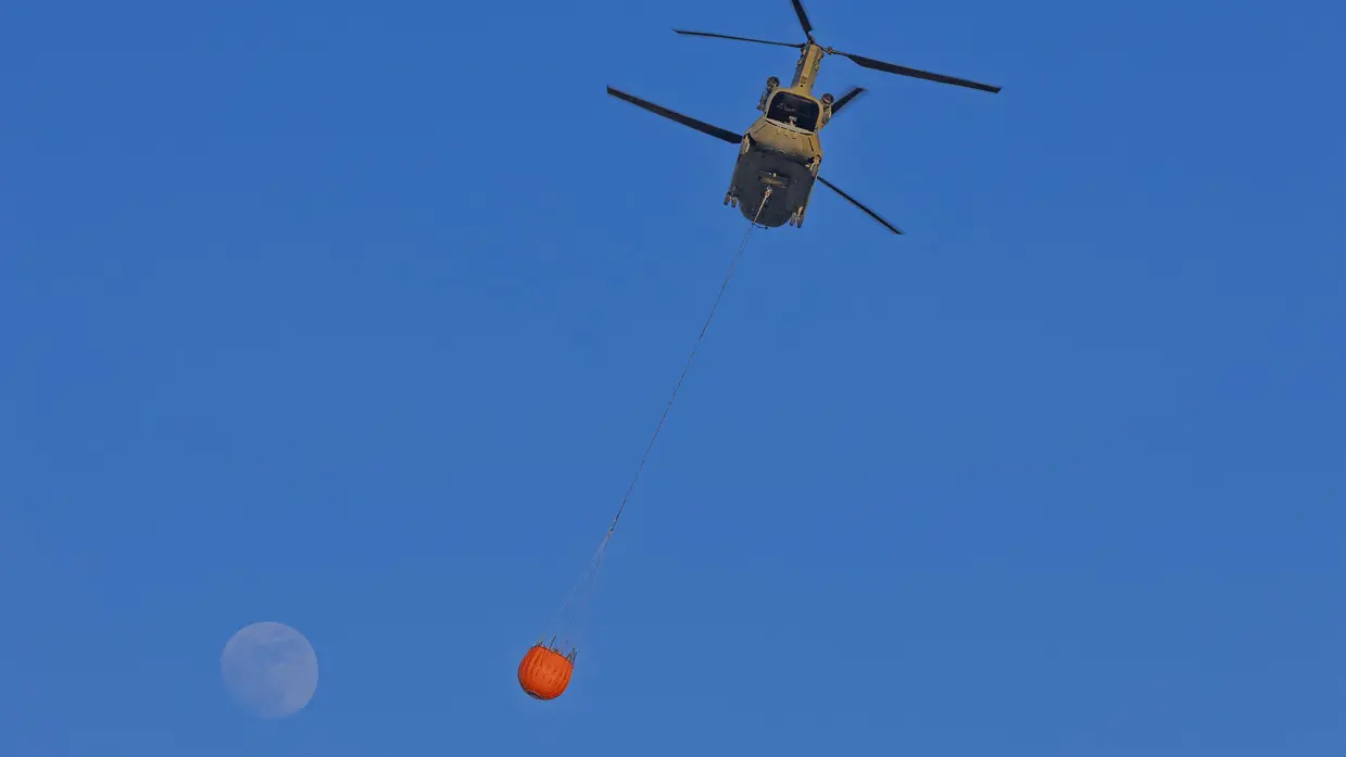Een helikopter hangt een grote oranje waterzak onder zich, met de volle maan op een diepblauwe hemel. 