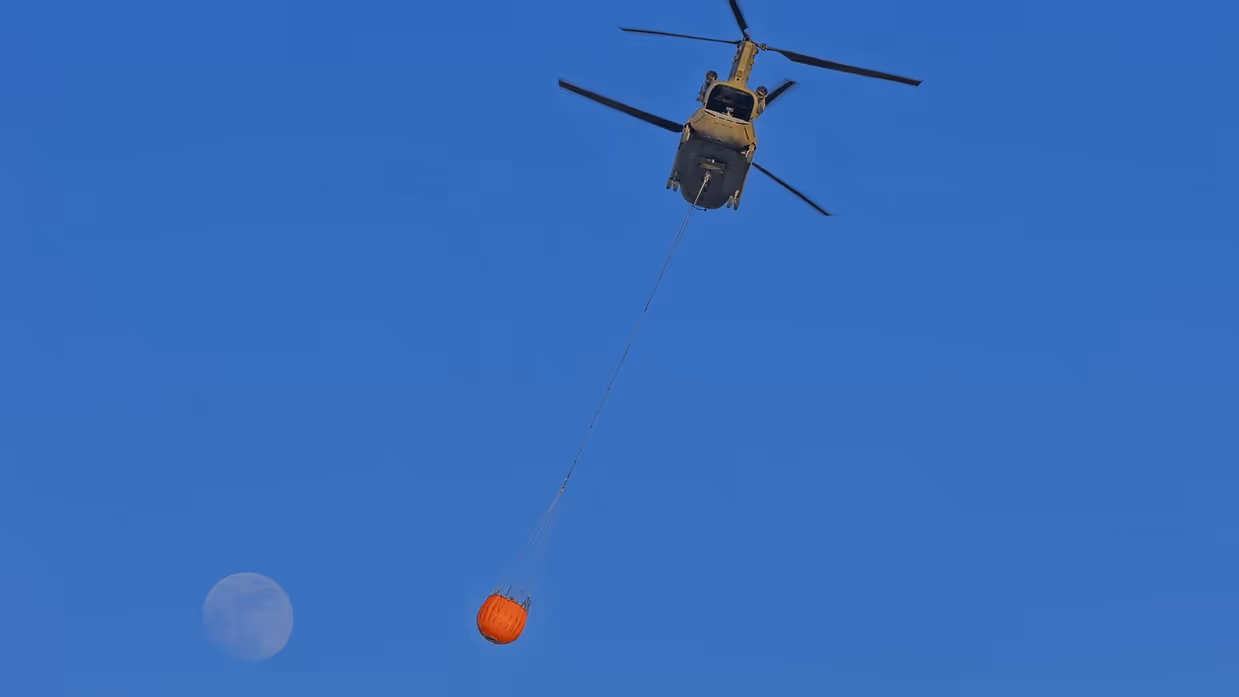 Een helikopter hangt een grote oranje waterzak onder zich, met de volle maan op een diepblauwe hemel. 