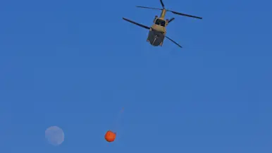 Een helikopter hangt een grote oranje waterzak onder zich, met de volle maan op een diepblauwe hemel. 
