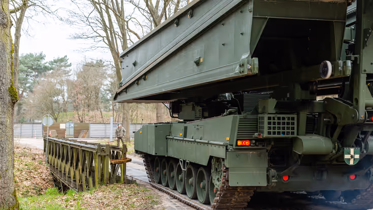 +alttekst: Een groene bruglegger legervoertuig met rupsbanden plaatst een zware metalen brug over een pad, omringd door bomen en een hek. 