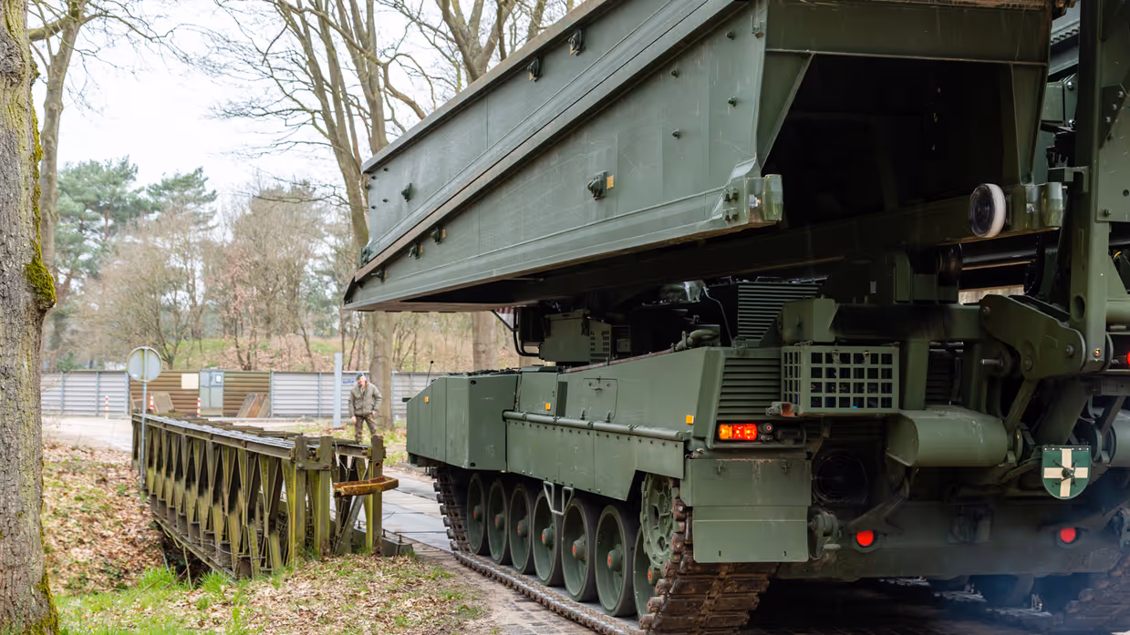 +alttekst: Een groene bruglegger legervoertuig met rupsbanden plaatst een zware metalen brug over een pad, omringd door bomen en een hek. 