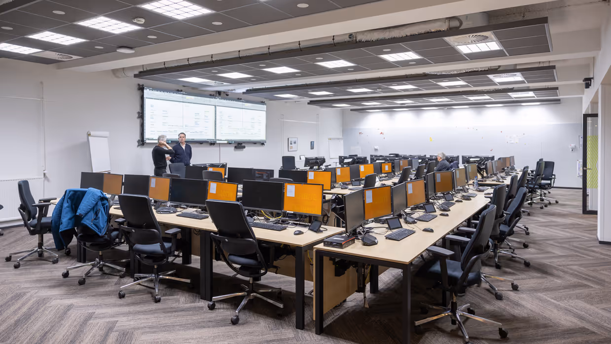 Een zaal met een dubbele, u-vormige bureauopstelling. Er zijn geen mensen aanwezig en alle computerschermen zijn oranje.