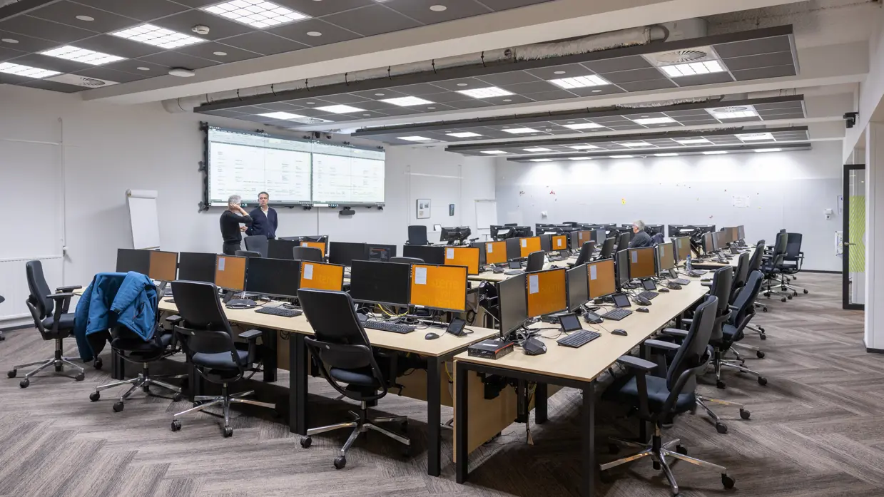 Een zaal met een dubbele, u-vormige bureauopstelling. Er zijn geen mensen aanwezig en alle computerschermen zijn oranje.