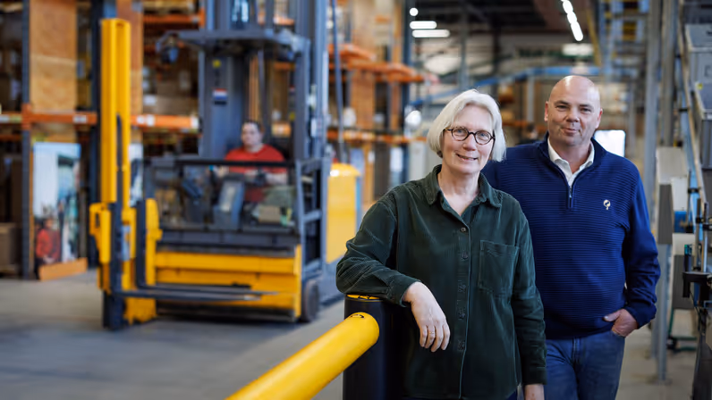 Portretfoto van Rick en Mieke in het distributiecentrum van het KPU-bedrijf. Op de achtergrond passeert een heftruck. 