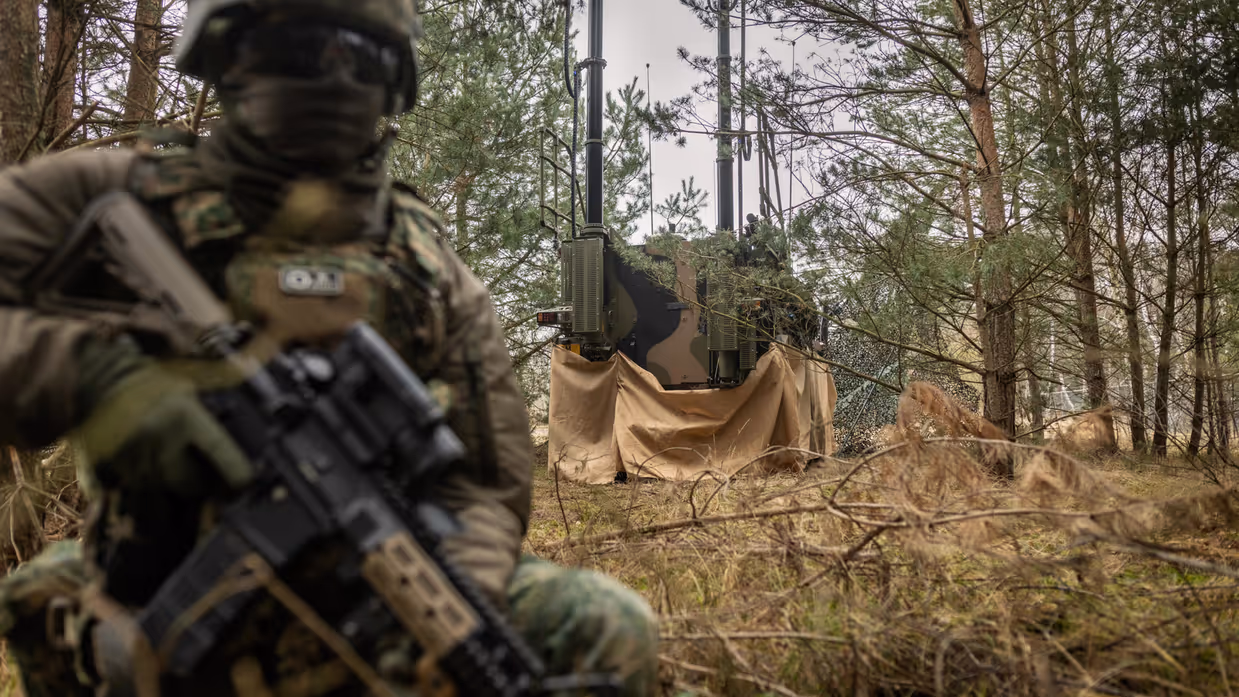 Een gecamoufleerde militair houdt op een knie gewapend de wacht aan de achterzijde van een Bushmaster, die in een groene strook van bos en struik geparkeerd staat. Op de Bushmaster zijn diverse antennes te zien. Foto rechts: Niels kijkt recht in de camera. Hij draagt een lichtgroene blouse en heeft kort donker haar. 