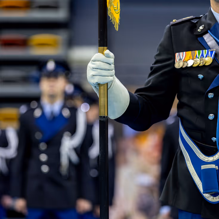 Marechaussee met vaandeel in de hand met op de achtergrond collega’s in de houding.