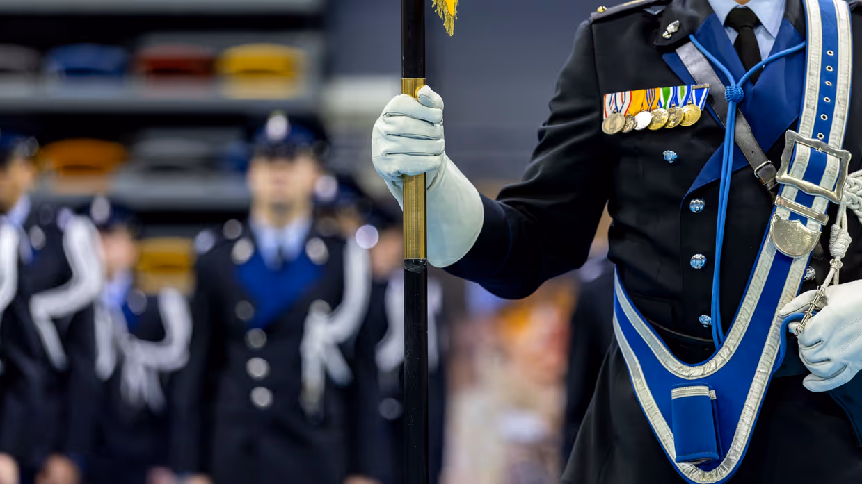 Marechaussee met vaandeel in de hand met op de achtergrond collega’s in de houding.