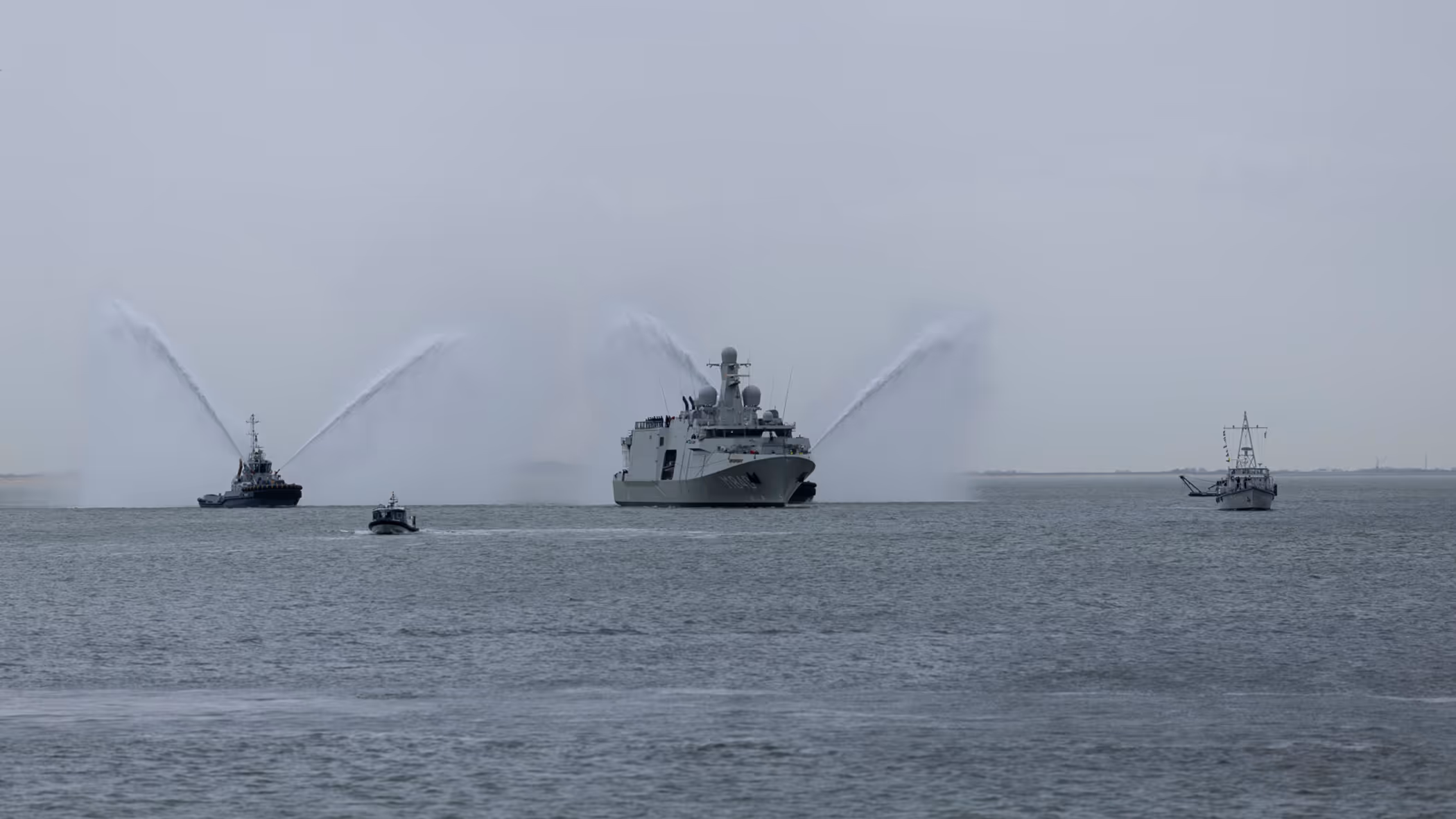 Marineschip Vlissingen op zee, daarnaast 2 sleepboten die met hun waterkanonnen spuiten. Het schip wordt voorgegaan door een kleine, klassieke mijnenveger.