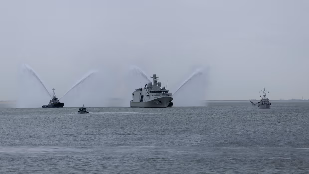 Marineschip de ‘Vlissingen’ op zee, daarnaast twee sleepboten die met hun waterkanonnen spuiten. Het schip wordt voorgegaan door een kleine mijnenveger. Op de tweede afbeelding hetzelfde schip, maar dan gezien langs een tweetal marineofficieren aan de wal. 