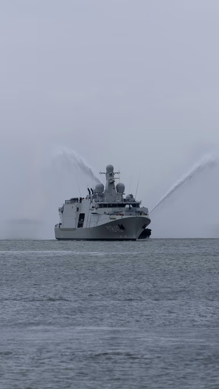 Marineschip Vlissingen op zee, daarnaast 2 sleepboten die met hun waterkanonnen spuiten. Het schip wordt voorgegaan door een kleine, klassieke mijnenveger.
